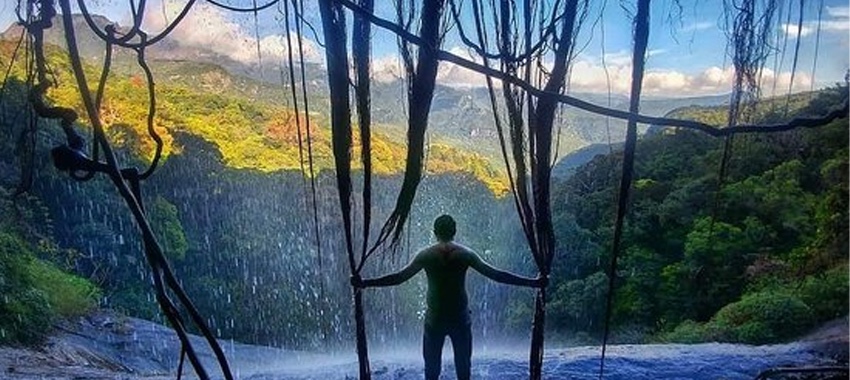 A traveller standing behind a waterfall overlooking the forested Central Highlands